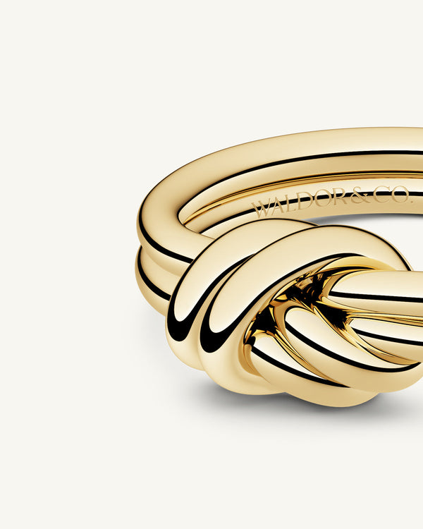 A polished stainless steel ring in 14k gold from Waldor & Co. One size. The model is Dual Knot Ring.